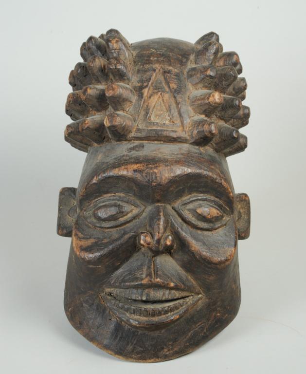 Bamun Helmet Mask: Carved wood mask with high, elaborate coiffure above a smiling face with round forehead, large eyes, full cheeks, naturalistic nose and wide smiling mouth with two rows of even teeth. Cameroon. Approx