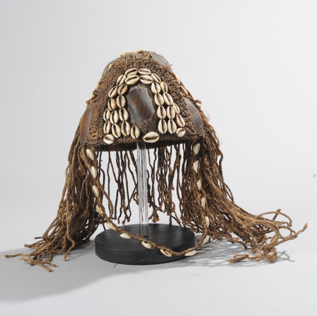 Lega Bwami Society Hat: A conical base of tightly woven cane and grasses, embellished with knotted string and shells. Braided raffia chin strap trimmed with cowries. Democratic Republic of Congo. On custom gallery stand. Hat