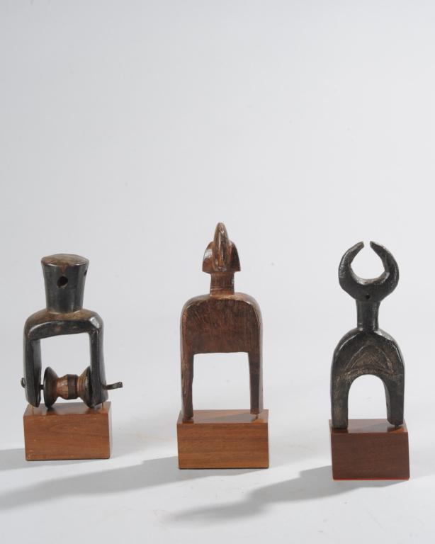 Three West African Heddle Pulleys: One with curved horns, perhaps an abstract buffalo, another roughly anthropomorphic, likely Bamana, the last with a thick, plug-shaped finial. All on custom gallery bases. Tallest (including base,) ap