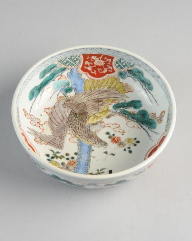 Antique Imari Bowl: The center with dramatic eagle swooping down towards a fenced garden with fir tree and flowers. Exterior with fir branches, butterflies and flowers between borders. Red and white shield shaped floral 