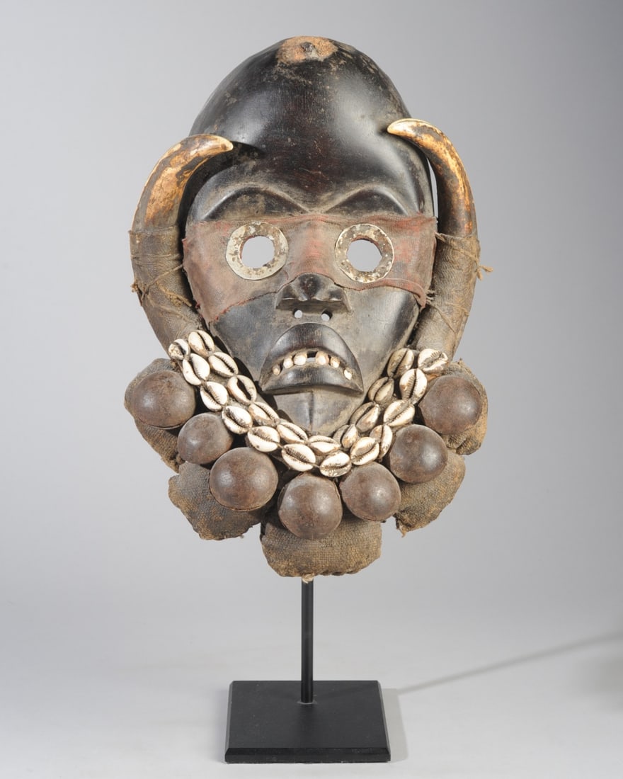 Dan Gunyege Mask: Carved wood mask with high round forehead, arched brows, the large round eyes encircled with tin, the mouth with corners drawn down and with a row of white teeth made of shell. The top of the brow has