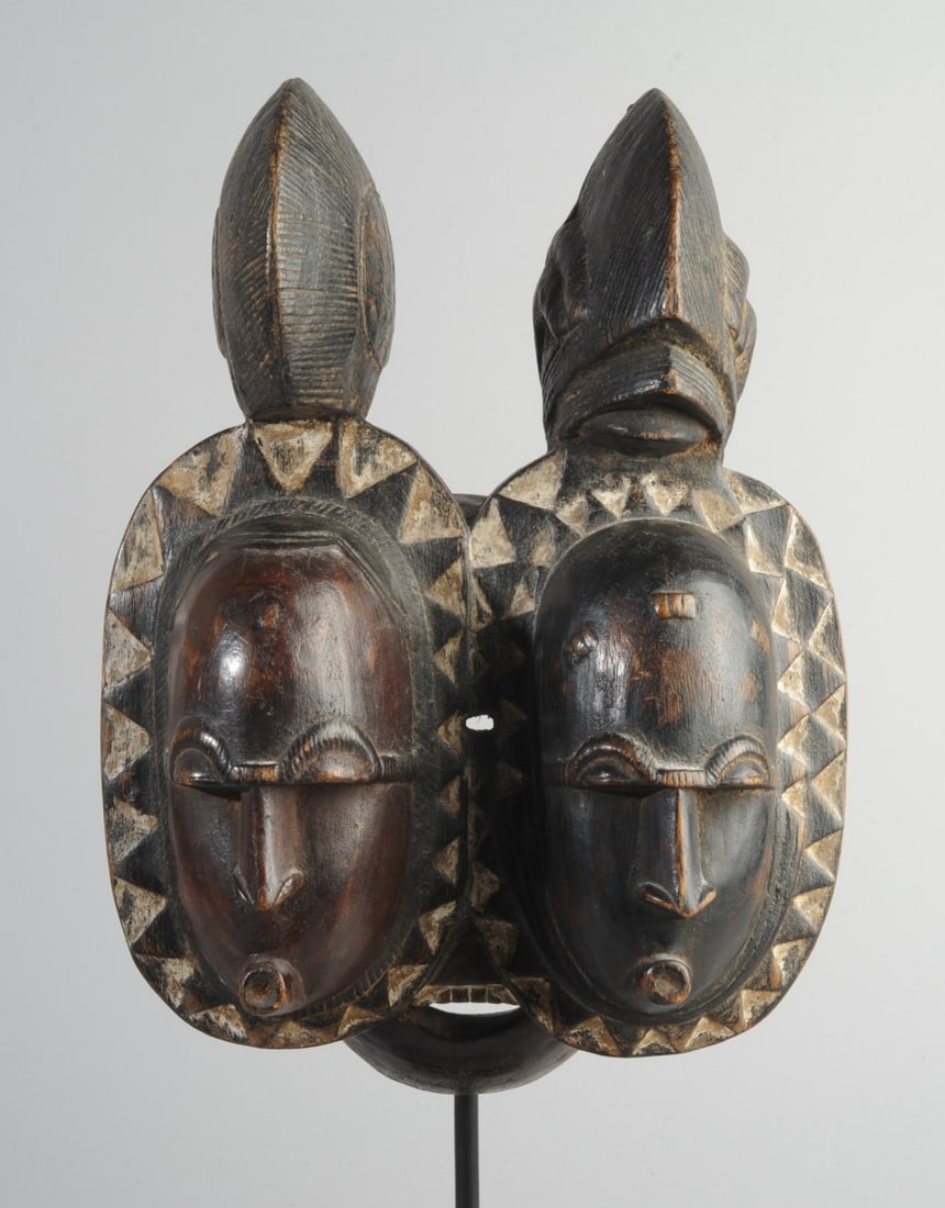 Baule Nda Mask, Ivory Coast: Carved wood double mask, the twin faces nearly identical but carved with subtle differences, both with elaborate carved topknots or headdresses, and both faces surrounded by saw-tooth borders. On cust
