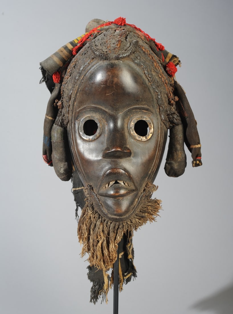 Dan Gunyege Mask w/ Accoutrements, Liberia: Carved wood Dan mask, the round eyes rimmed with metal, and with small metal teeth. The elaborate coiffure is formed of braided and twisted raffia heavily packed with clay or similar material. The col