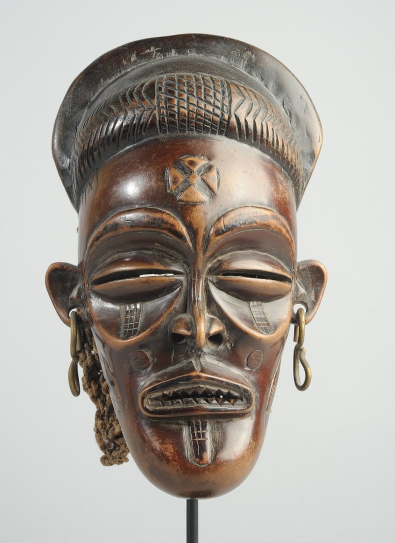 Chokwe Mwana Pwo Mask, Angola: Smallish carved wood mask with typical cruciform mark on brow, stylized tears, facial scarification, and filed teeth. With metal earrings and rope netting hood. On custom gallery stand. Mask, approx. 