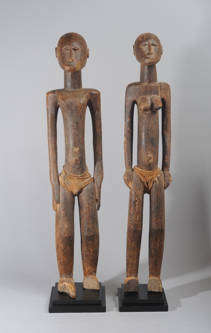 Large Lobi Couple Figures, Burkina Faso: Finely carved wooden standing male and female figures, their posture and facial features expressing strength and serenity. Slightly encrusted surface. Each wearing a cloth garment. Both with losses to