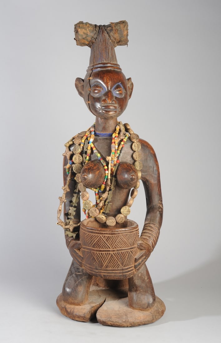 Yoruba 'Olumeye' Bowl Bearing Figure, Nigeria: Kneeling female shrine figure with high, conical coiffure or headdress, an offering bowl held in her hands. Blue pigment around eyes. Embellished with fabric, and with trade beads and metal beads. Dee