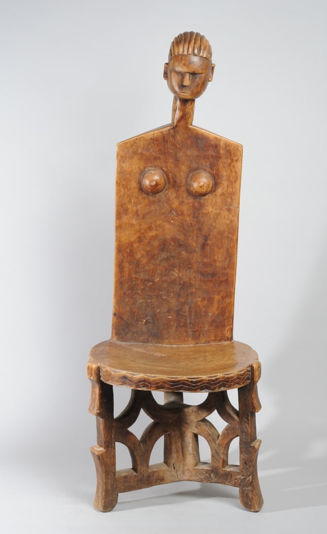 Kaguru Chair, Tanzania: The high, anthropomorphic back topped by head with swept back coiffure, deep-set eyes, minimalist features, on long neck above plank back with round breasts, the round seat with wavy lines carved alon