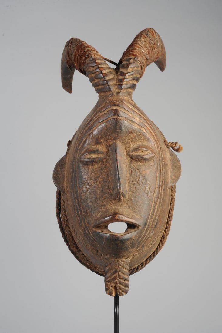 Mossi Horned Mask, Burkina Faso: Mossi mask with blend of human and animal features, with curved goat-like horns partially wrapped in strips of animal hide with residual reddish fur, the flat facial plain with coffee bean eyes, long 