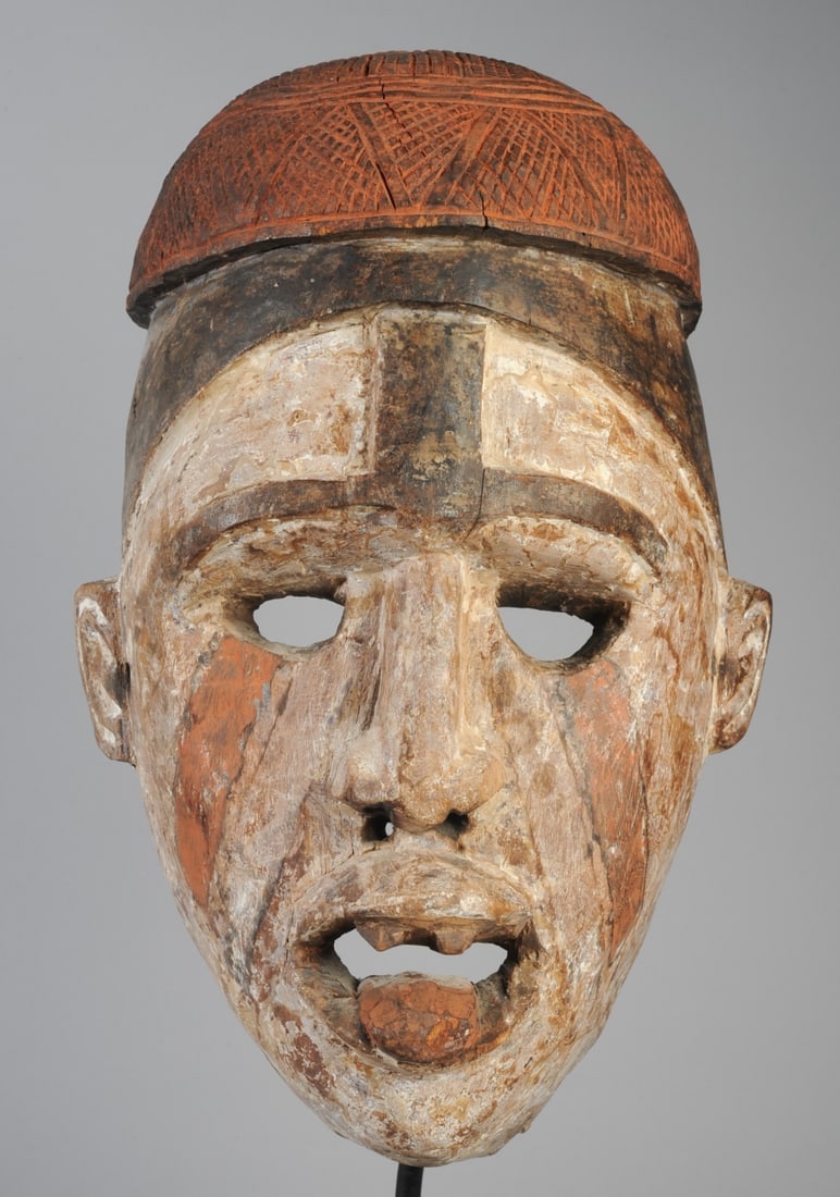 Bakongo / Yombe Helmet Mask, D. R. Congo: Anthropomorphic mask with red cap, whitened face with naturalistic features, hollow eyes, mouth open with teeth showing and tongue protruding. Red-orange diagonals below eyes. On custom gallery stand.