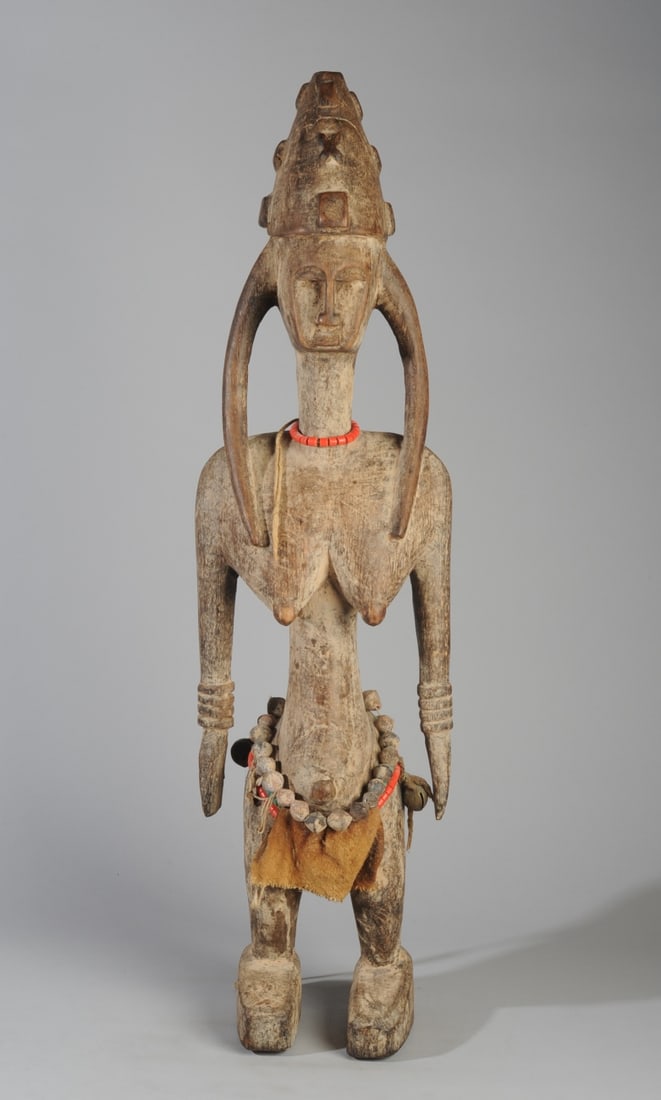 Bamana or Marka Figure, Mali or Guinea: Standing female figure wearing conical cap with relief carved symbols over hair which descends in three long plaits. The wedge shaped head has a flat facial plane with deep set eyes, long straight nos