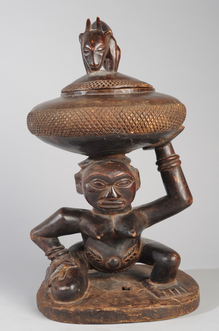 Luba Caryatid Bowl Bearer Figure: Crouching female figure with one knee on the ground, one in squat posture, supporting a round lidded bowl on the top of her head and with one raised hand. the lid of the bowl with finial in the form o