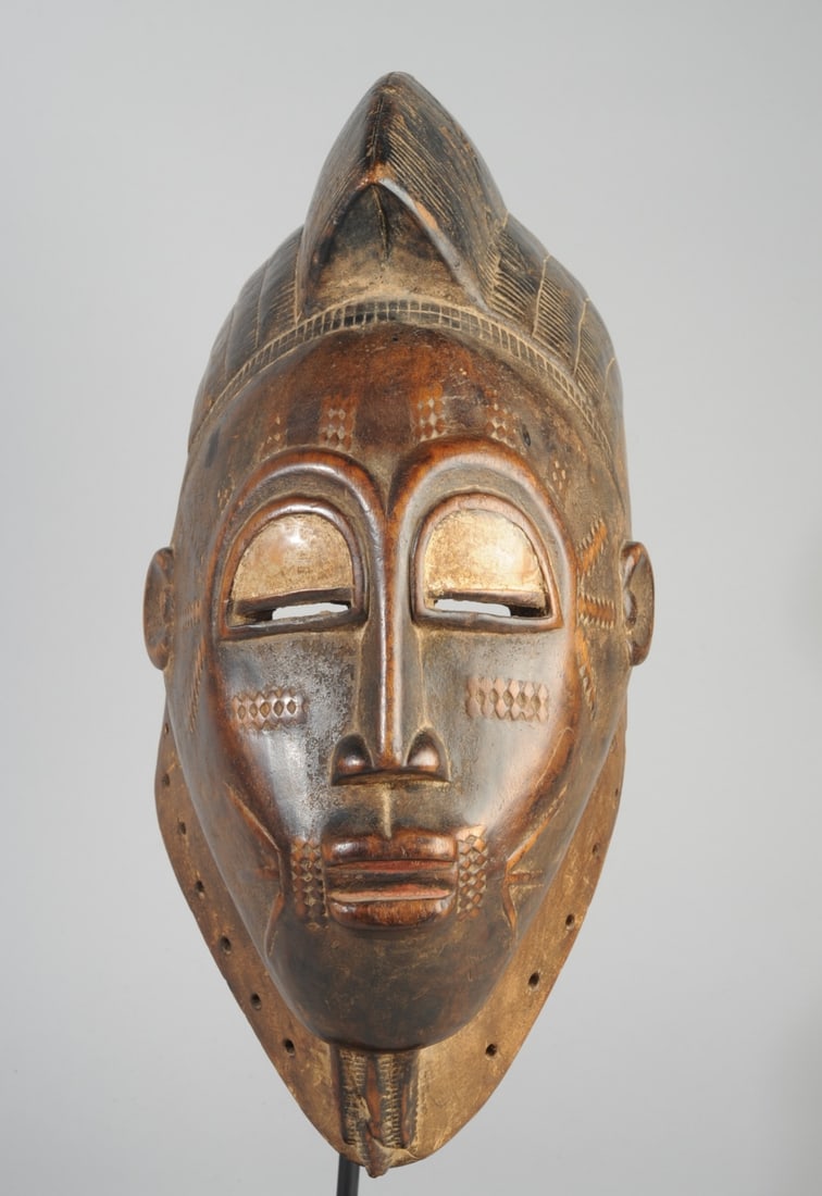 Guro Mask, Ivory Coast: Helmet mask, the coiffure with high central lobe , the heart shaped face with high arched brow, slender nose, small ears, the parted lips plump. Many relief carved keloids at brow, temples, cheeks, an