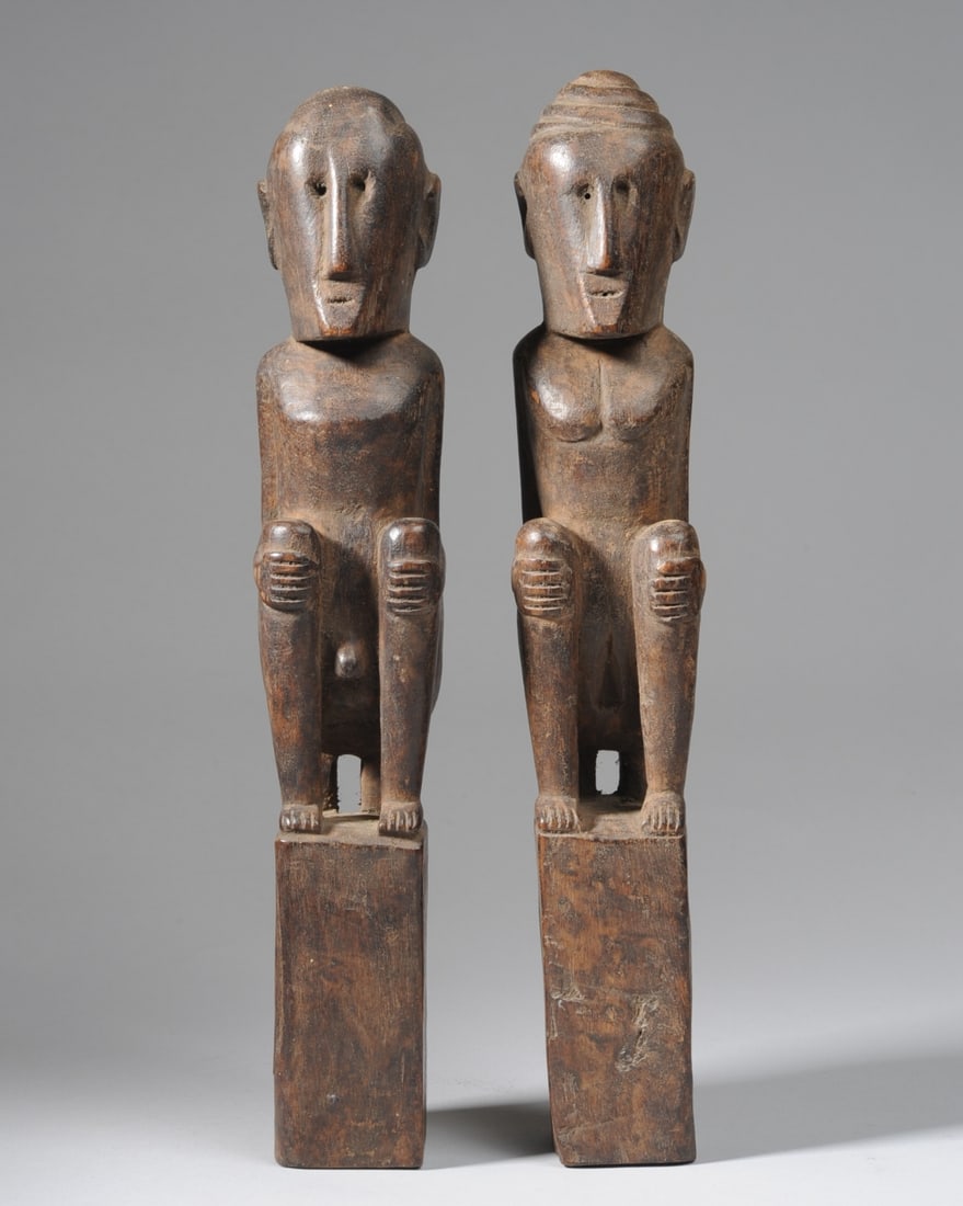 Pair Flores Ana Deo Ancestor Figures, Indonesia: Pair male and female "ana deo" ancestor figures, carved squatting with hands grasping upraised knees. Both with narrow faces tapering towards chin and minimalist facial features. The male has a cap-li