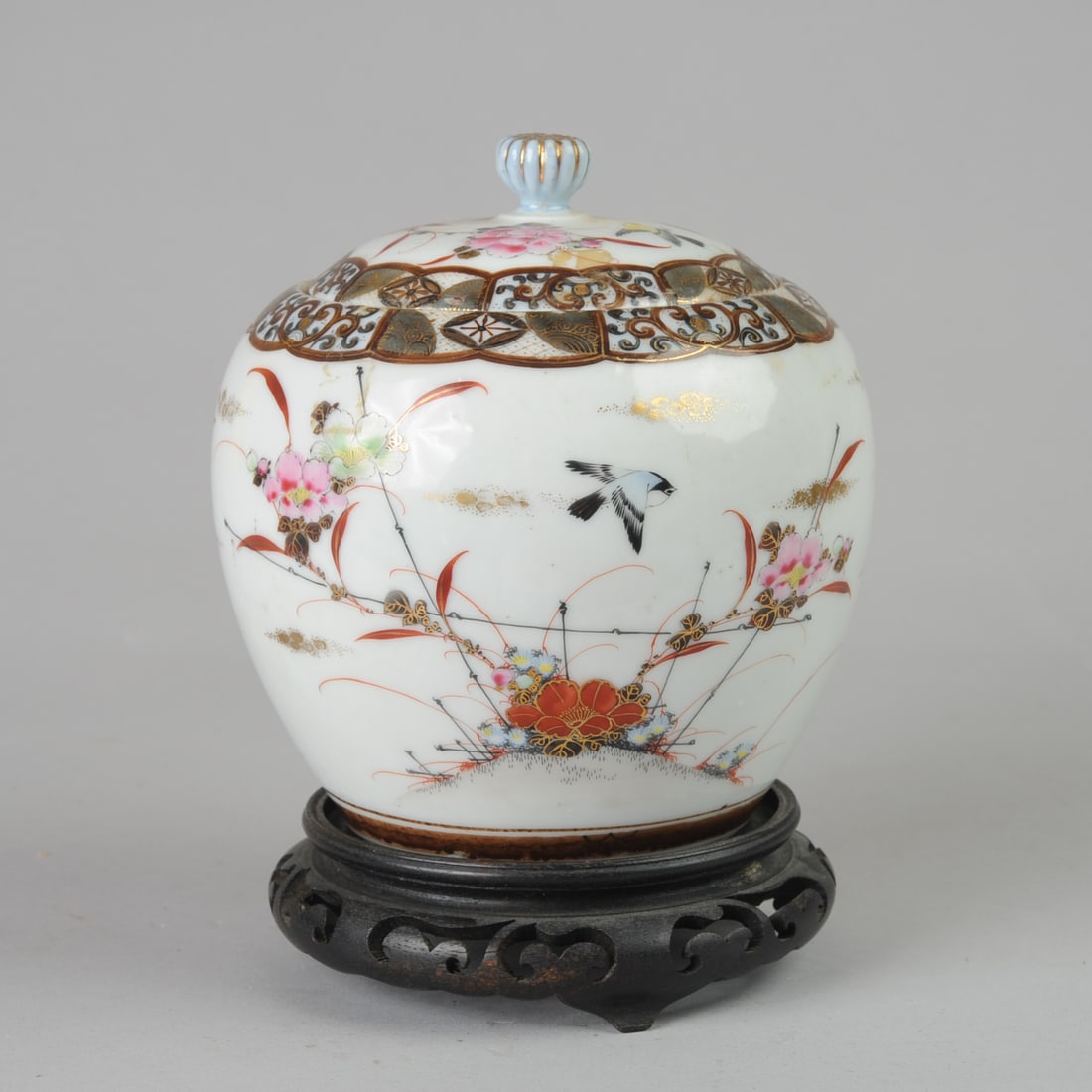 Japanese Porcelain Meiji Kutani Jar and Cover: Antique Japanese Kutani porcelain jar with birds and flowers in enamels and gilt, with chrysanthemum bud knop. Likely a tobacco jar. With carved wood stand. Jar (not including stand,) approx. 6" H x 6