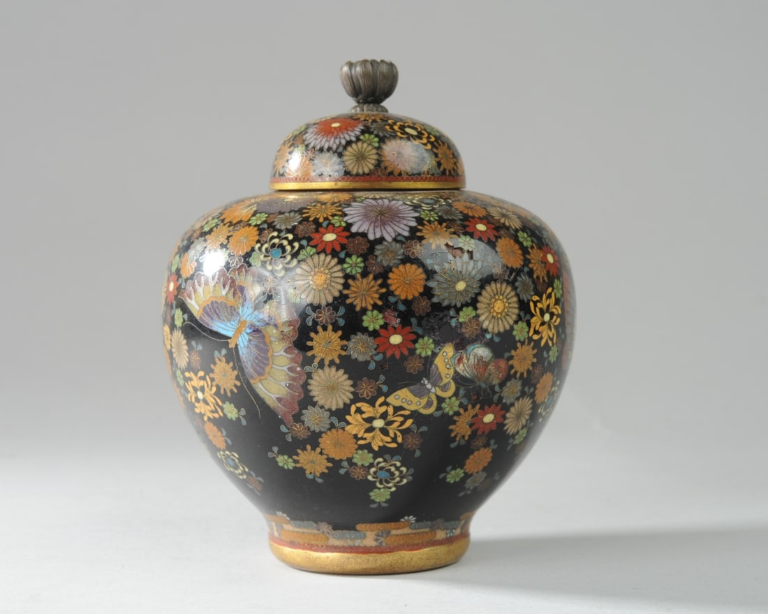 Japanese Cloisonne Meiji Period Jar & Cover: Fine and detailed cloisonne flowers and butterflies on black ground with narrow red border. Chrysanthemum form knop. 3/4" shallow dent with cracks to enamel on one side. Approx. 5 1/4" H., 4" D. Seaml