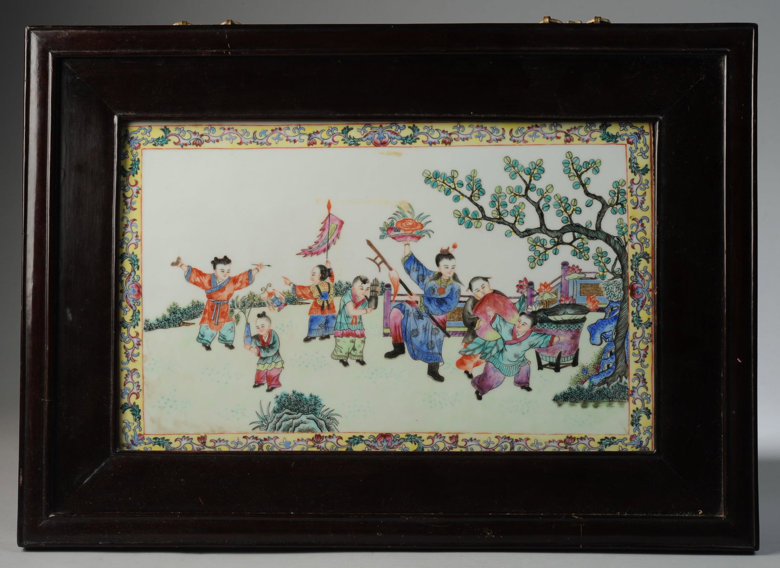 Chinese Porcelain Plaque: Framed porcelain plaque with figures of children on a terrace garden celebrating with a giant peach, a banner, an ink stone and brush, a flute, a lantern, and other auspicious objects in famille rose 