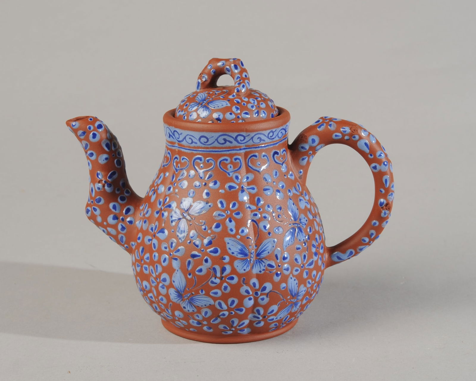 Blue Enameled Yixing Teapot: Red zisha clay with flowers, butterflies and borders in light and cobalt blue enamel. lobed body with twisted branch handle, finial and spout. Impressed mark to base. No chips, cracks, hairlines or re