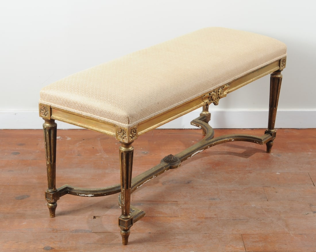 Louis XVI Style Upholstered Giltwood Bench: Fluted, tapered legs topped by rosette squares and joined by curved X stretcher with central medallion. Apron with carved floral sprays on front and back. Champagne colored fabric with raised diamond