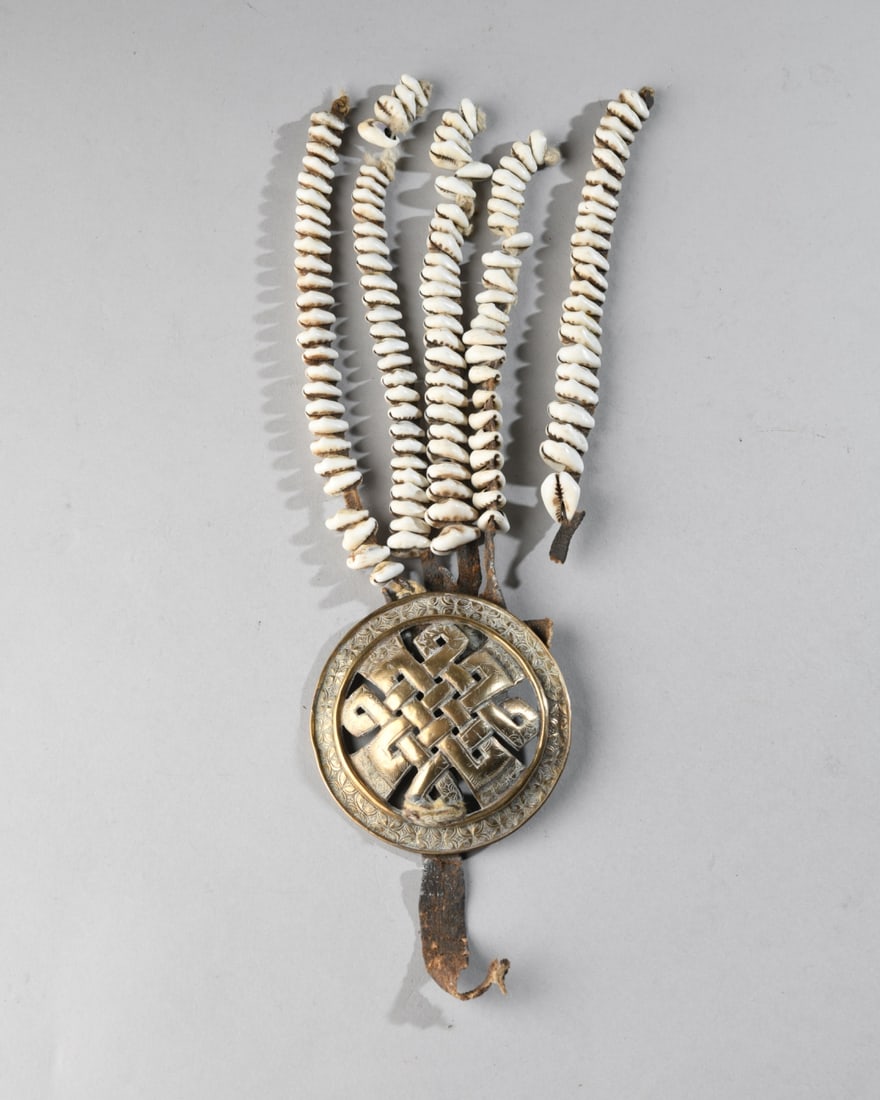 Ladakh Dodchas Woman's Waist Ornament: The traditional woman's belt or waist ornament with large, chased brass disk with openwork interlacing knot, from which hang four leather strips strung with cowrie shells. One strip detached, likely