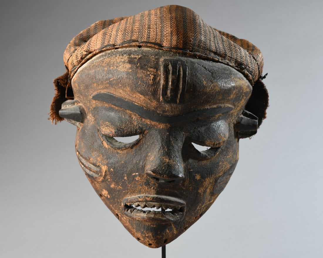 Pende Mbuya Mask, Ex-Alfred Scheinberg: Carved dark wood mask with tightly woven raffia headdress. Pende tribe, Zaire (now Democratic Republic of Congo.) On gallery stand. Mask, excluding stand, approx. 10" x 9". (Provenance: Purchased