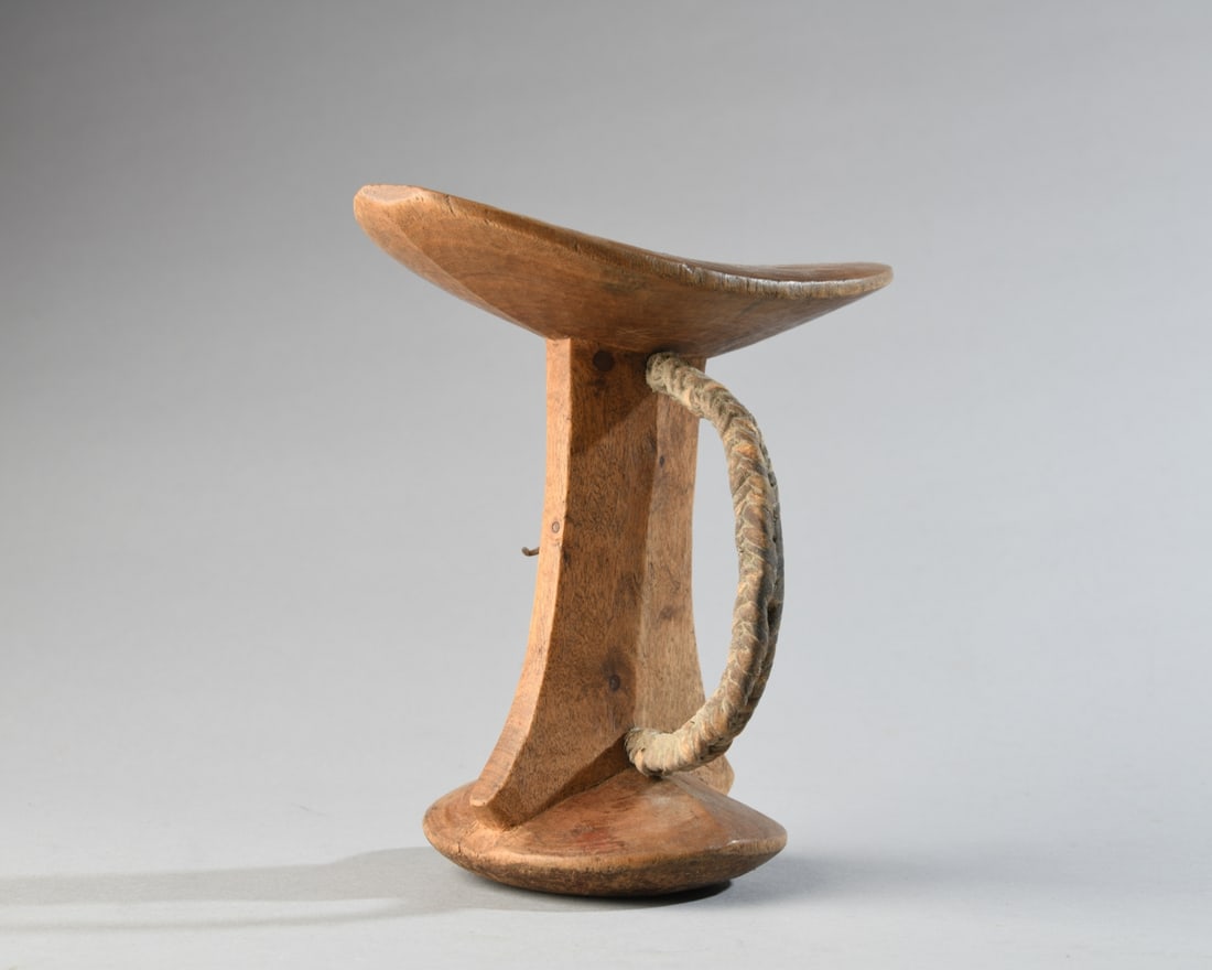 Masaai Headrest: Carved hardwood headrest, the oval top plate flattened on two sides, with flaring pillar, round base, and with plaited hide handle. Approx. 6 1/2" x 3 3/4" x 6 1/2" H. Our primary recommendation is