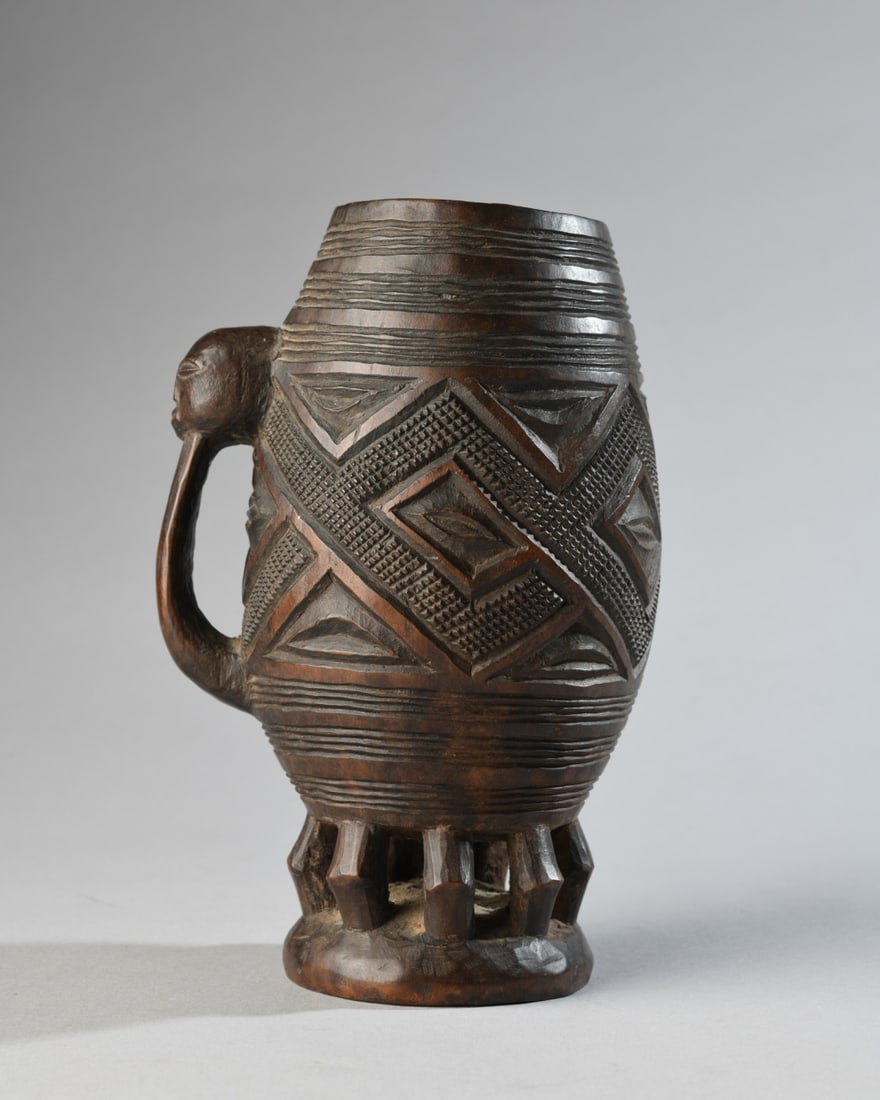 Kuba Palm Wine Cup, Ex-Christie's: Barrel-form cup on openwork base, the circumference with geometric carving of interlocking diamonds with central almond-shaped elements, the loop handle with thumb rest in the form of a human face.