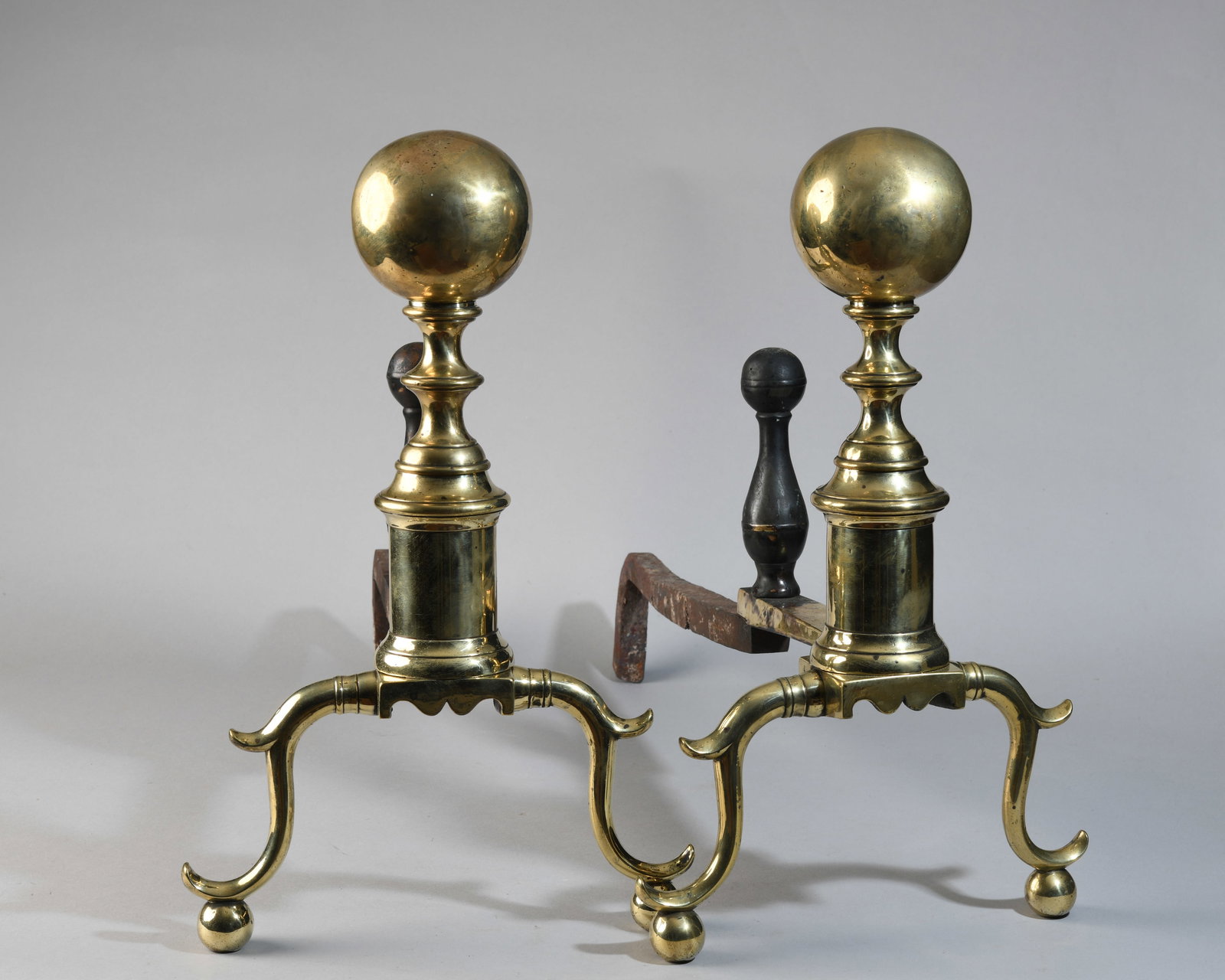 Pair of English Georgian Brass Andirons: Each with spherical finial above a turned shaft and square plinth, raised on three cabriole legs with pad feet, joined to wrought iron billets; 18th century with later elements; surfaces with tarnish,