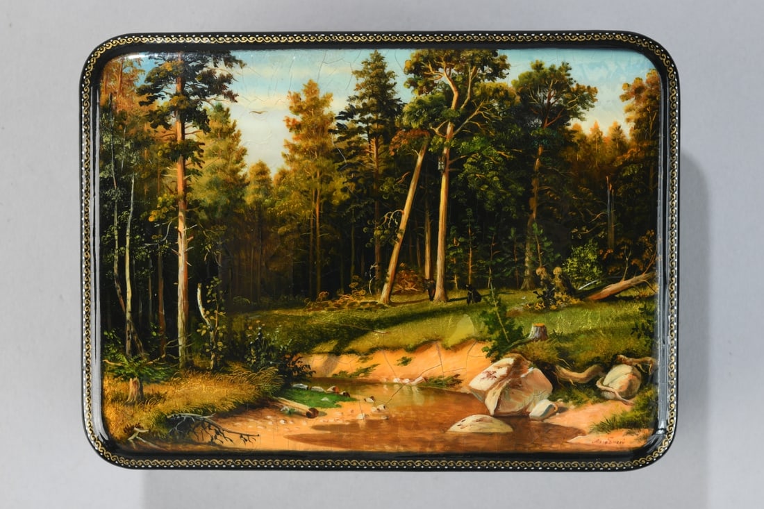 Lacquered Russian Box with Painted Landscape: Rectangular black lacquered box with hinged lid, the top painted with a forest landscape scene, bordered by a gilt scrollwork band, red interior, 20th century, surface with craquelure, minor wear. Dim