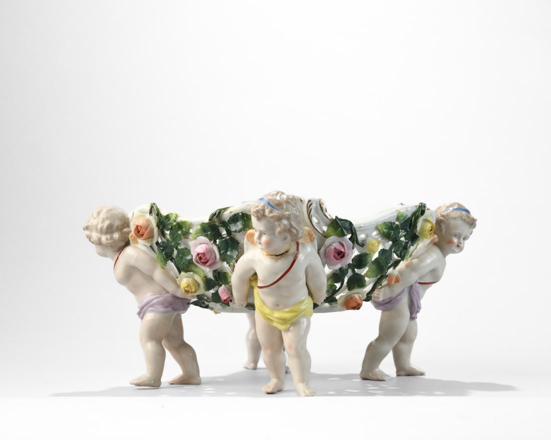 Continental Porcelain Figural Centerpiece Bowl: Glazed porcelain, late 19th to early 20th century, likely German or Austrian, modeled as a reticulated oval basket supported by three putti and applied with floral garlands, with polychrome enamel