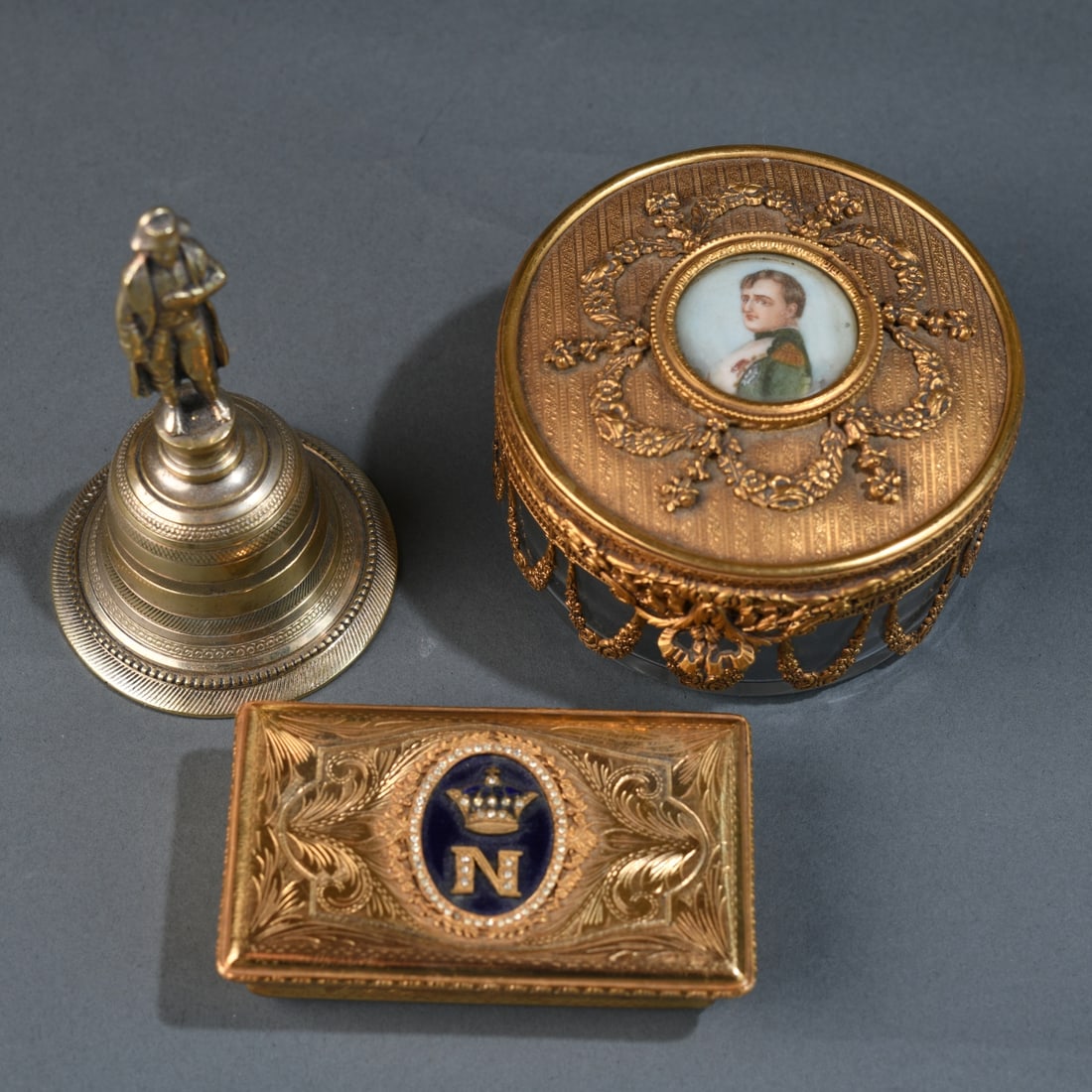Three Napoleonic Style Dresser Articles: Including a gilt metal and glass casket with portrait miniature of Napoleon, swag-mounted sides and star-cut base; a rectangular gilt metal box with blue enamel crowned “N”; and a small figura