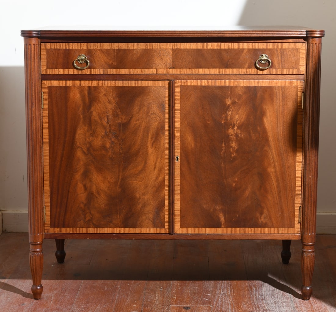 20th C. Hepplewhite Style Server: Shaped top with reeded edge, single dovetailed drawer with brass pulls over double doors flanked by reeded columns on turned feet. Dimensions approx. 34 3/4" H x 37 3/4" W x 19 1/4" D. Our primary