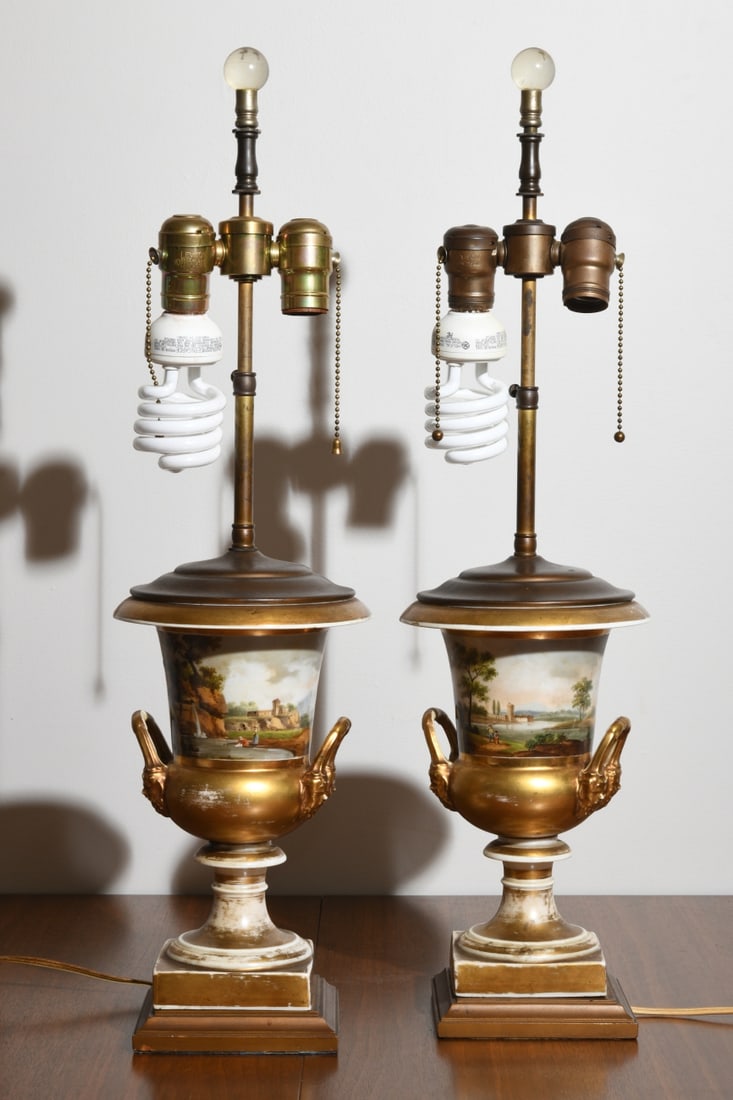 Pair of Painted Porcelain Table Lamps: Pair of porcelain urn-form table lamps with parcel gilt decoration and hand painted continuous pastoral landscapes, mounted with scrolling ram’s head style handles and square bases. Electrified and 