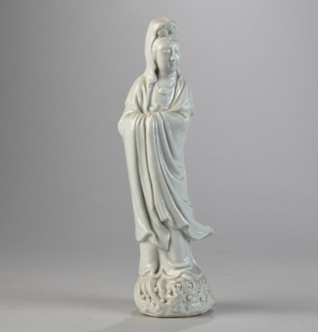 Chinese Porcelain Figure of Guanyin: Chinese blanc-de-chine style porcelain figure of Guanyin, modeled standing in flowing robes with hands concealed within sleeves, raised on a molded wave base; covered in a glossy white glaze. Undersid