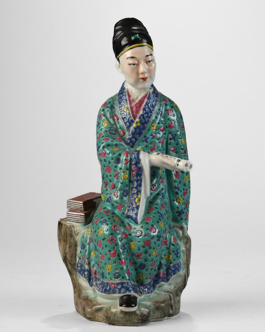 Chinese Porcelain Scholar Figure: Modeled as a seated male figure holding a scroll, wearing a green ground robe densely decorated with polychrome floral roundels and patterned borders in famille verte style enamels, the figure seated 
