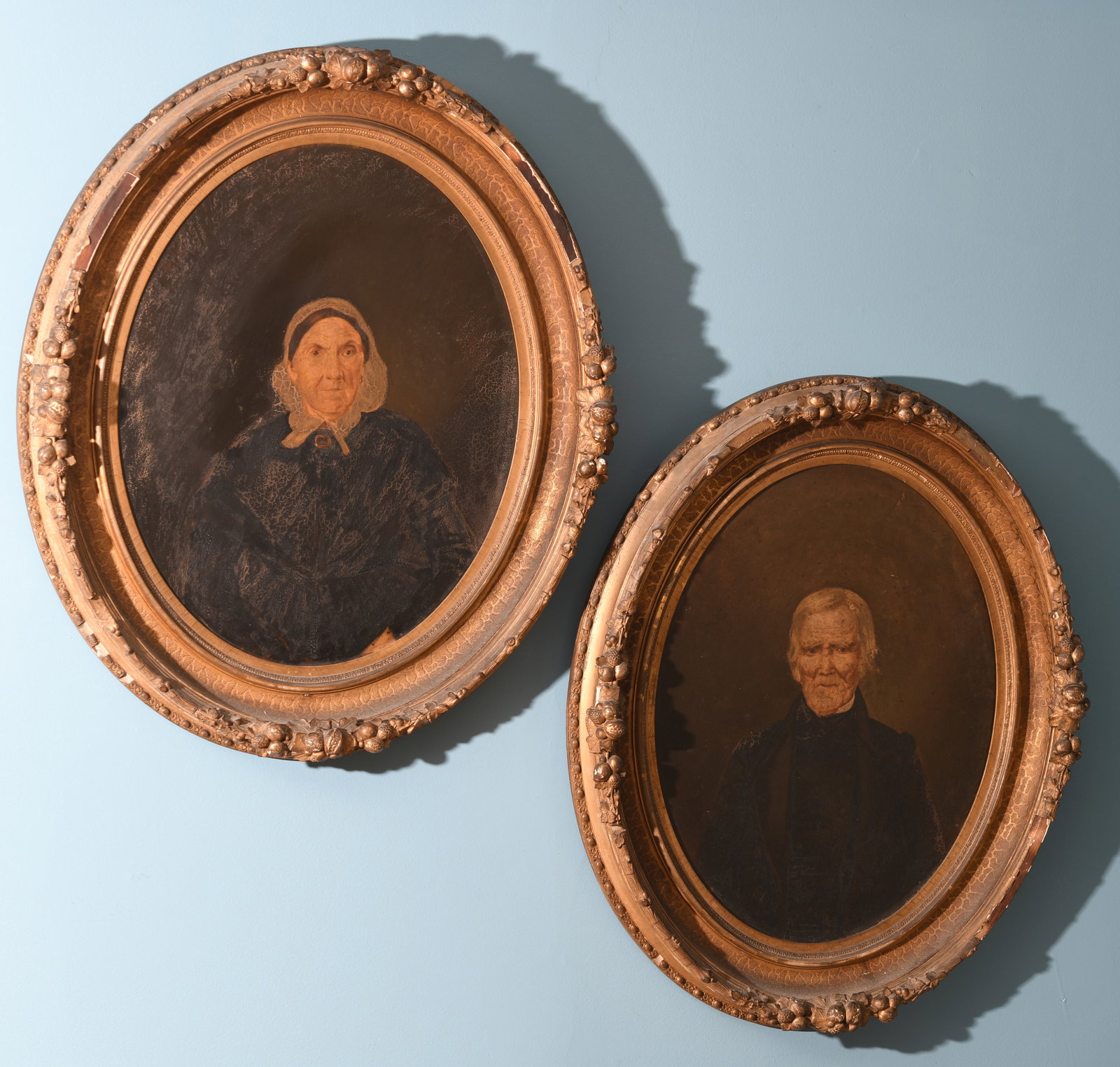 Pair 19th C. American Folk Portraits: Oil on board depicting an elderly woman in bonnet and dark dress and an elderly gentleman in black coat, both within matching oval gilt composition frames with applied fruit and foliate cresting, unsi