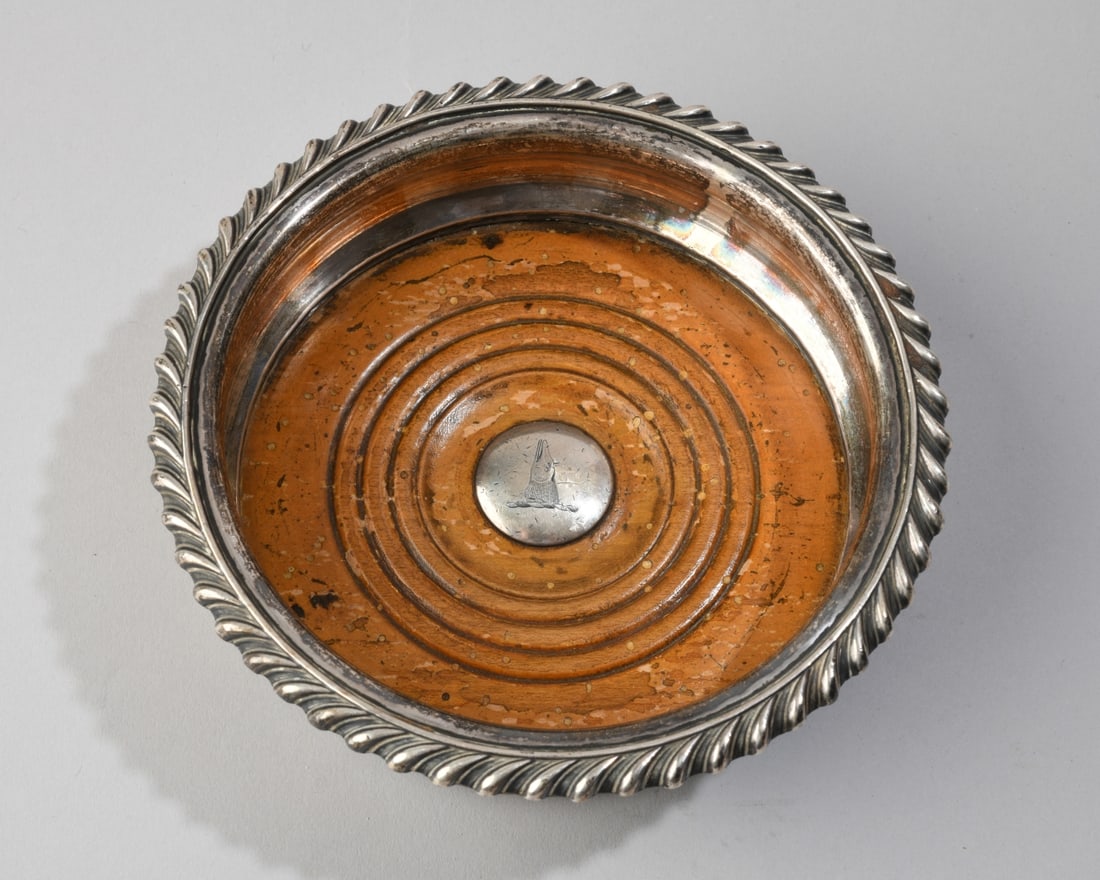 Antique English Silverplate Wine Coaster: Rolled edge rim over turned wood base inset with engraved crest depicting a pike or fish head. No visible maker's marks. Dia: approx. 6", H: approx. 1 7/8". Our primary recommendation is to arrange