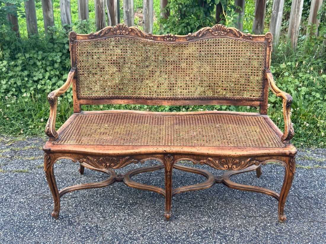 French Louis XV Style Carved Caned Settee: Probably late 19th century, shaped crest rail decorated by shell, scroll, and lattice-carved motifs above a continuous cane back, open scrolling arms, caned seat, cabriole legs and shaped stretcher