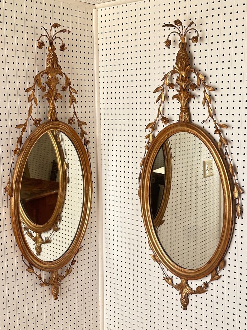 Pair of Gilt Oval Wall Mirrors: 20th century, with elaborate openwork crest and pineapple finial, foliate swags, one shows a crack / split in wood frame. Approx. 48 1/2" x 18 1/2". Our primary recommendation is to arrange shipping