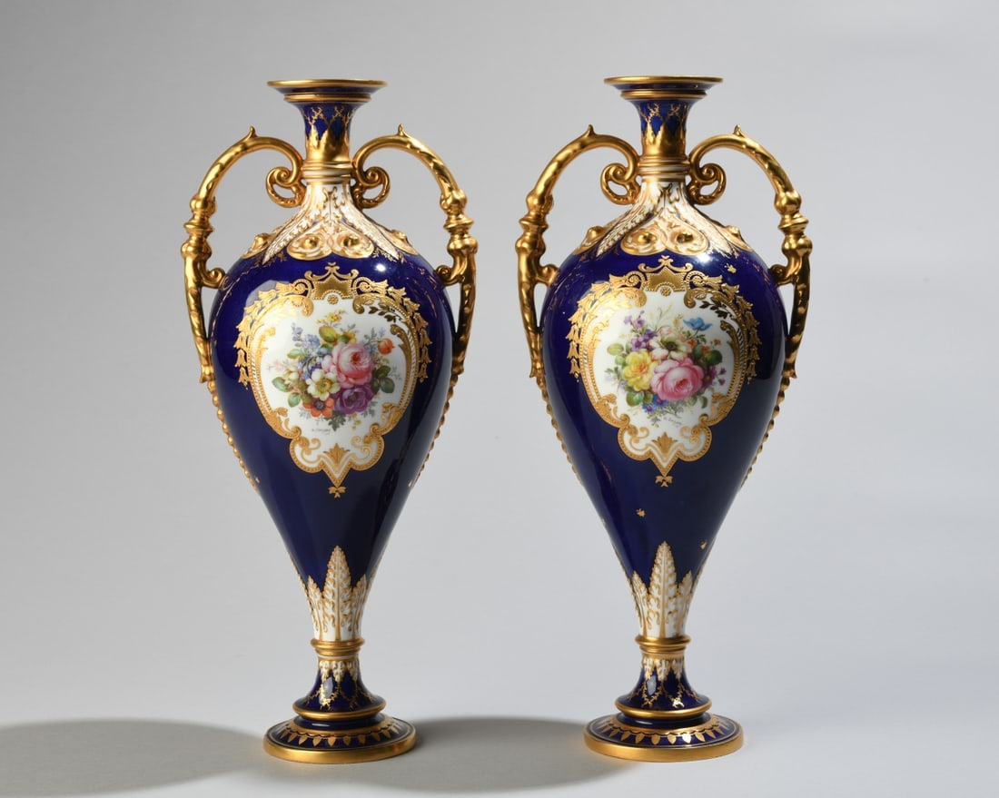Pair of Royal Crown Derby Cabinet Vases: With double handles, cobalt blue ground, richly embellished with raised and gilt scrollwork and foliate ornament, each side centered by an oval reserve painted with floral bouquets. Marked to