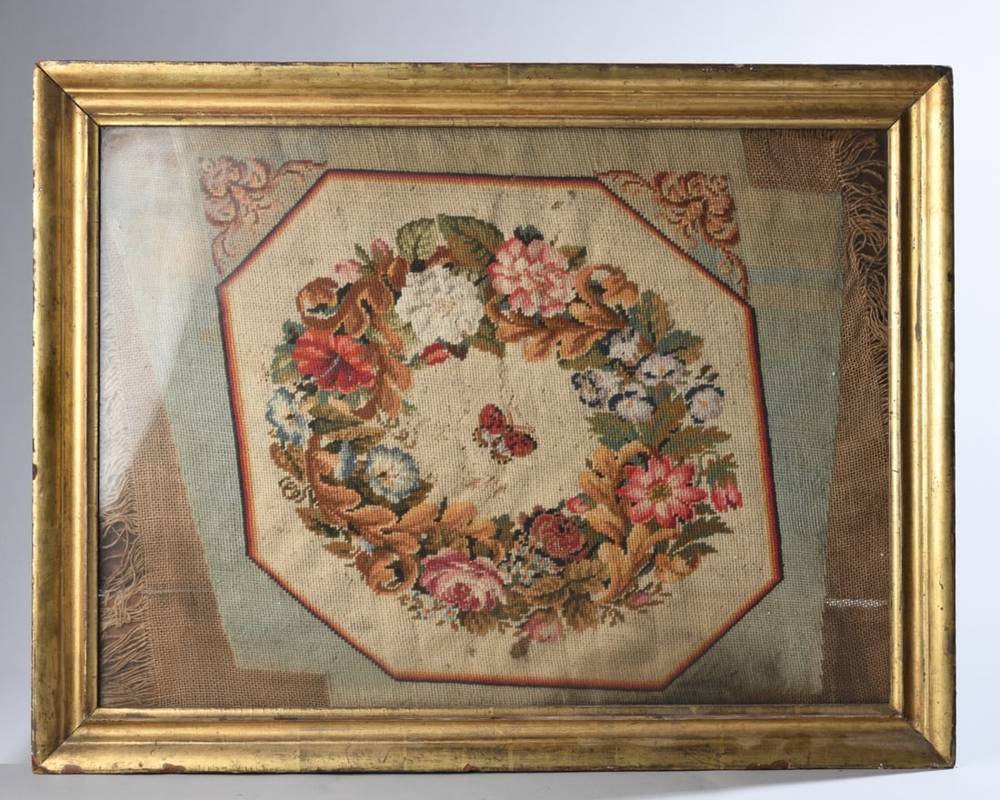 Framed Needlework: Featuring floral wreath and butterfly design on canvas. Overall fading, discoloration, and stitch wear; losses and wear to frame. Sight dimensions approx. 24 1/4" x 17 3/4". Frame dimensions approx.