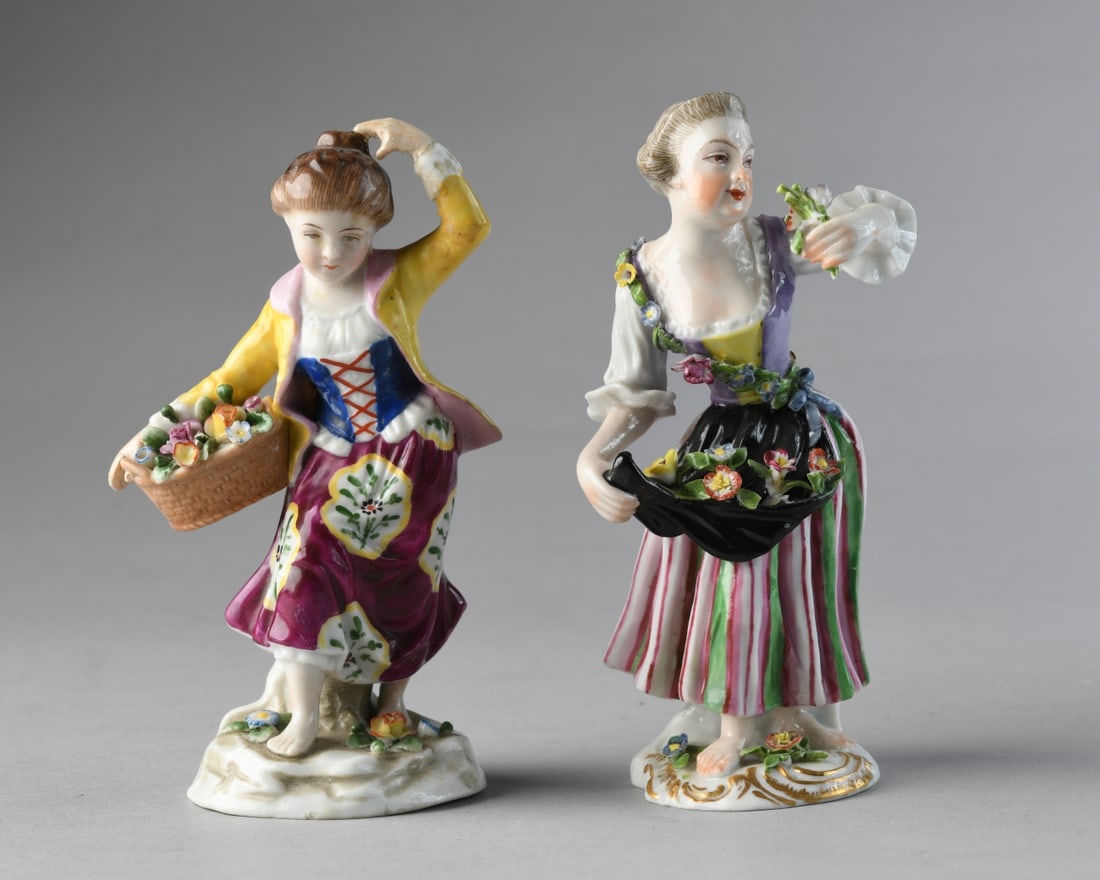 Pair of Continental Porcelain Figures: Two hand painted porcelain figures depicting women holding floral baskets, each with applied flowers and molded bases. One with anchor mark to backside, the other with gilt SS mark to underside. One