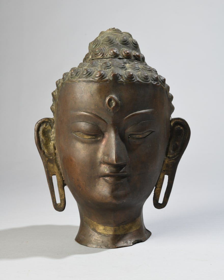 Southeast Asian Bronze Buddha Head: Cast bronze head of Buddha with downcast eyes, elongated earlobes, urna to forehead, and tightly curled hair beneath a domed ushnisha, on integral flared neck base. Surface wear, chip / loss to base,