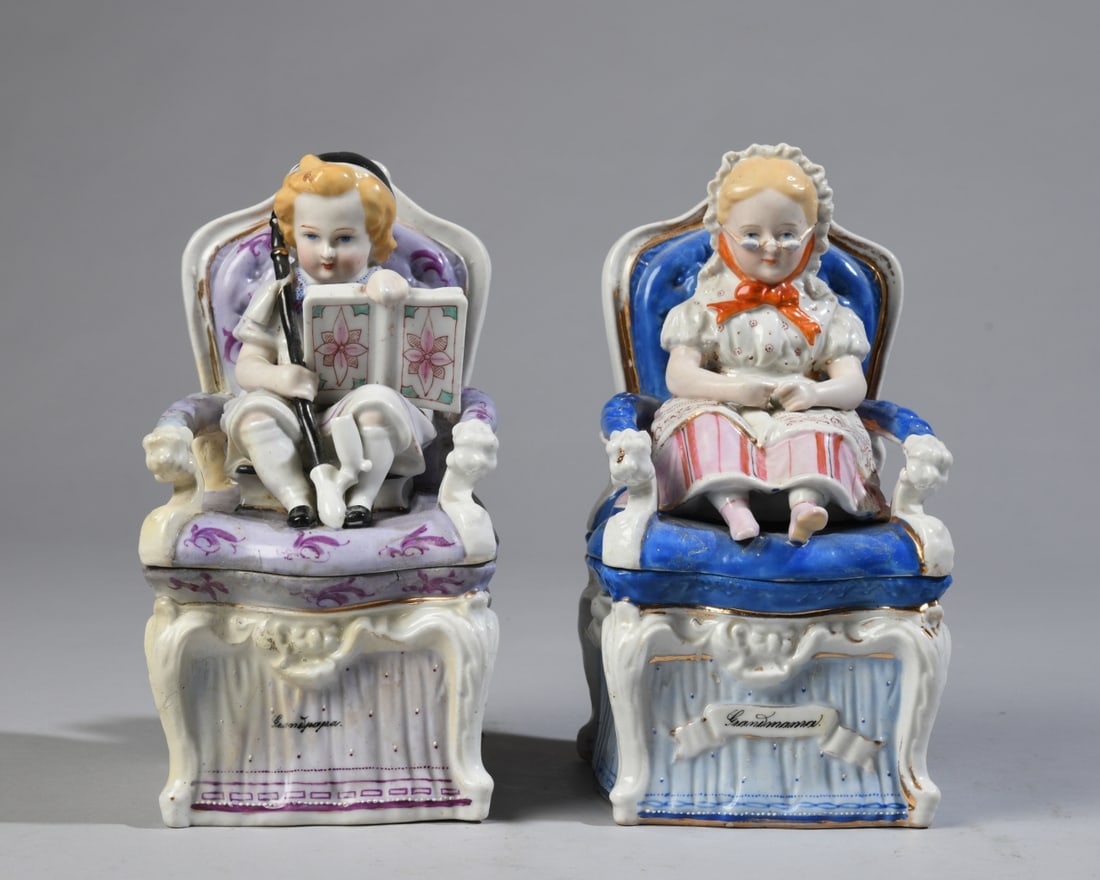 Pair German Porcelain Figural Tobacco Jars: Depicting children seated in armchairs, labeled "Grandpapa!" and "Grandmamma!" to the fronts. Each separates at the chair seat to form a covered container; impressed model numbers to underside.