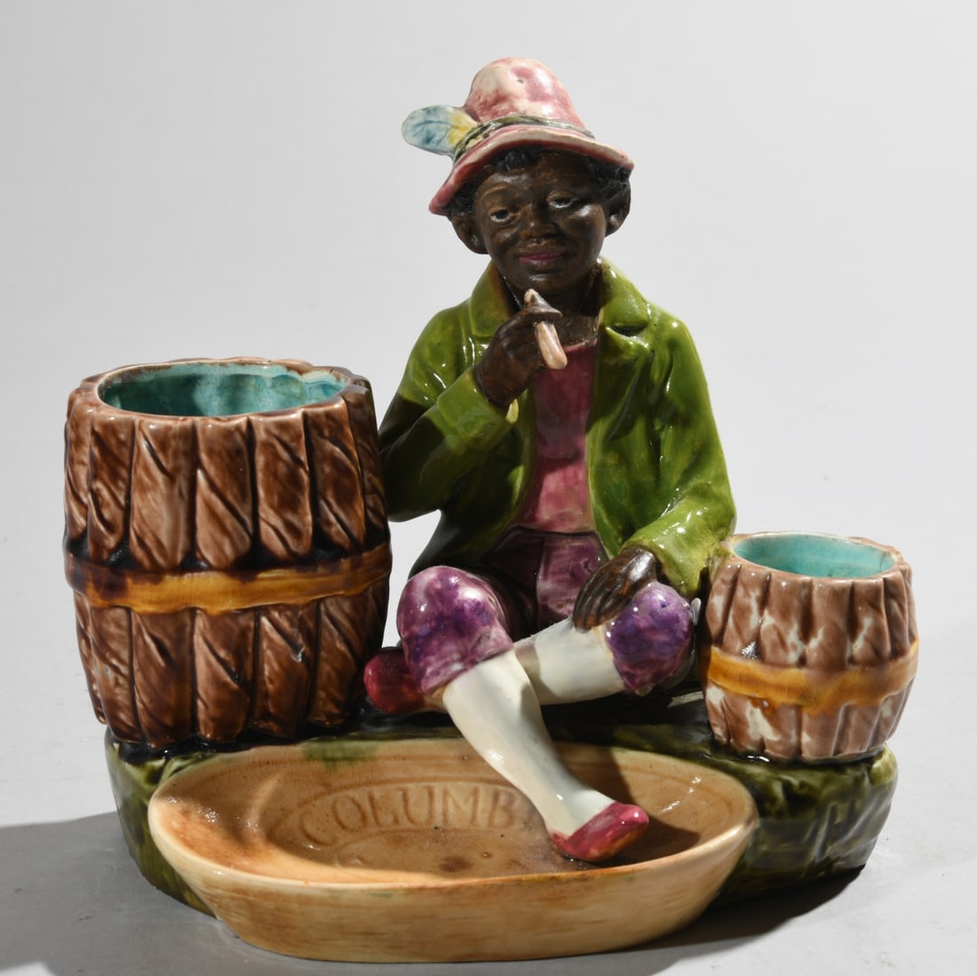 Continental Majolica Figural Smoking Set: Modeled with a seated figure smoking a cigar between two barrel form receptacles, with integral oval ashtray impressed 'COLUMBIA' and an indistinct second word, with ribbed match striker to the