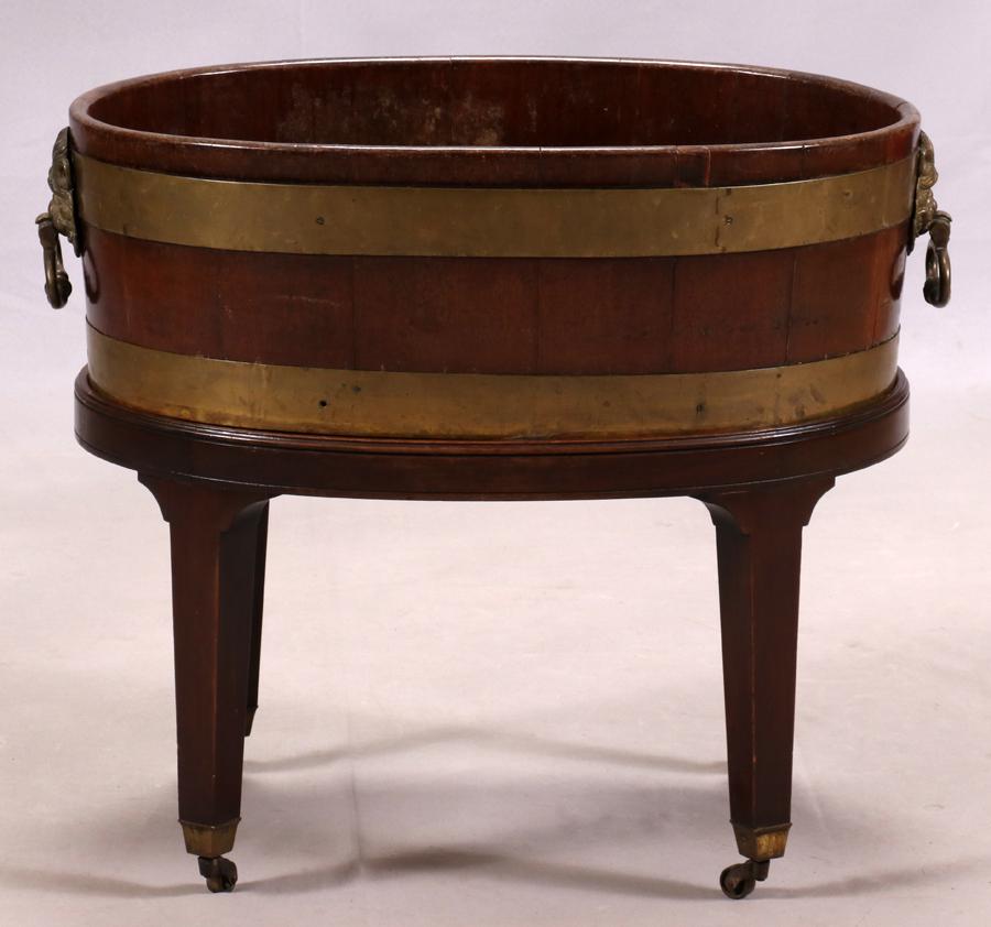 GEORGE III BRASS-BOUND MAHOGANY WINE COOLER: GEORGE III BRASS-BOUND MAHOGANY WINE COOLER, C. 1770, H 21 1/2", W 26" In the manner of Thomas Chippendale, oval with lion mask and ring handles, on straight legs with casters.