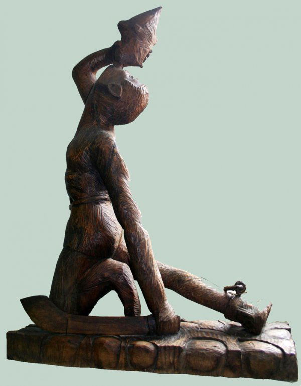 HAITIAN CARVED WOOD FIGURE, H 53", W 14", L 35": HAITIAN CARVED WOOD FIGURE, H 53", W 14", L 35", 'DESSOLINE':Figure depicted is kneeling and is blowing into a conch shell. He has a shackle on his right foot and holds a machete in his left hand.