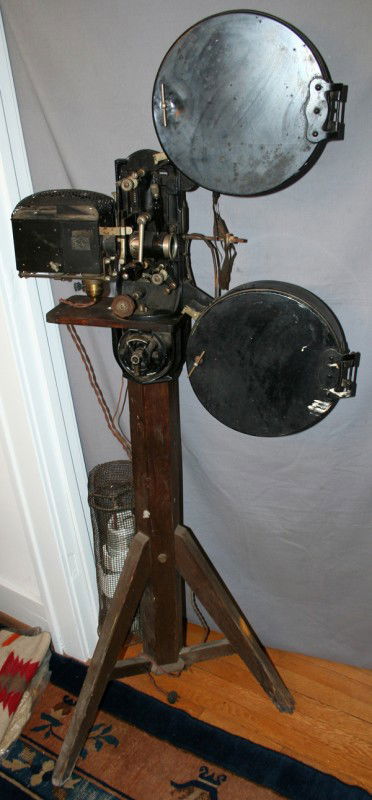EARLY SILENT ERA 35MM MOVIE PROJECTOR, C1920,: EARLY SILENT ERA 35MM MOVIE PROJECTOR, C1920, H 65", L 26":hand crank operated, used 35 mm acetate film; having a wood pedestal and tripod legs; two attached metal film reel cans; metal plate with ser