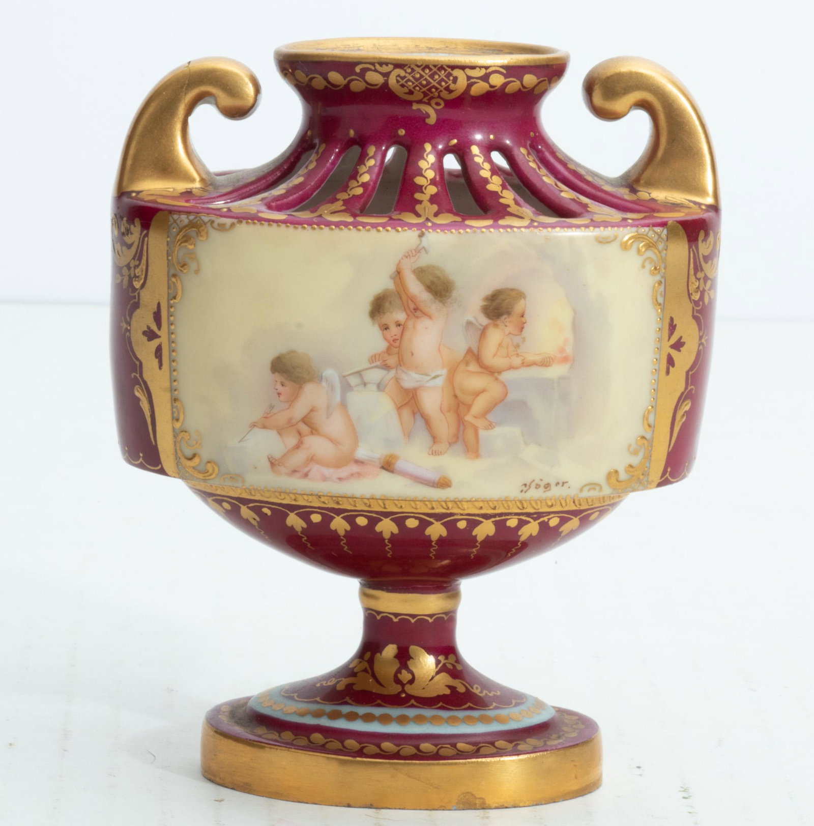 ROYAL VIENNA, AUSTRIA, PORCELAIN URN, C 1880 H 4 3/4": ROYAL VIENNA, AUSTRIA, PORCELAIN URN, C 1880 H 4 3/4" Having a deep red body and 2 open gold handles. Hand painted vignette depicting playful cherubs, Signed Roger. On a short stem pedestal. Signed wi