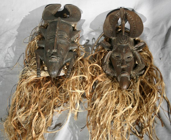 CARVED AFRCIAN CEREMONIAL MASKS, 2 PCS.,: CARVED AFRCIAN CEREMONIAL MASKS, 2 PCS., H 16" & 17":having integral carved horn shape headresses, one with carved leg forms and other with boar tusk forms all carved from the wood; each has straw sus
