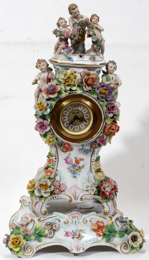DRESDEN PORCELAIN CLOCK ON STAND, CARL THIEME,: DRESDEN PORCELAIN CLOCK ON STAND, CARL THIEME, EARLY 20TH C., H 15 1/2", W 9":The porcelain clock case surmounted by five putti, adorned with flowers, and raised on a separate base; the clock and base